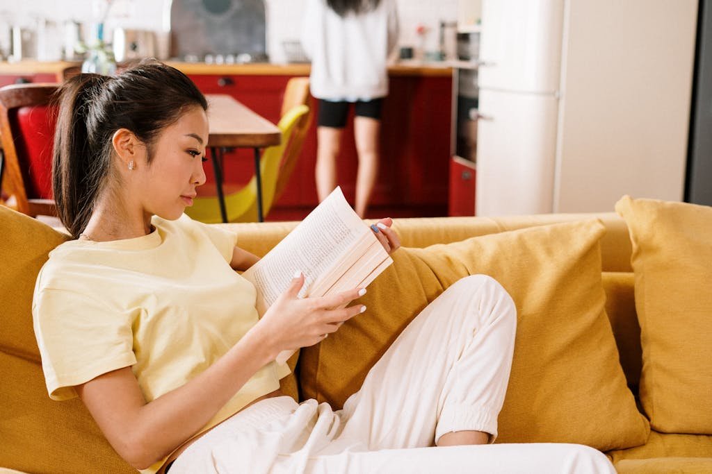 Asian woman relaxing on a cozy yellow sofa, reading a book in a modern home interior.