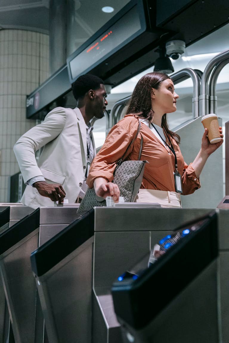 Black man and Caucasian woman rush through a subway turnstile holding a coffee and a laptop case.