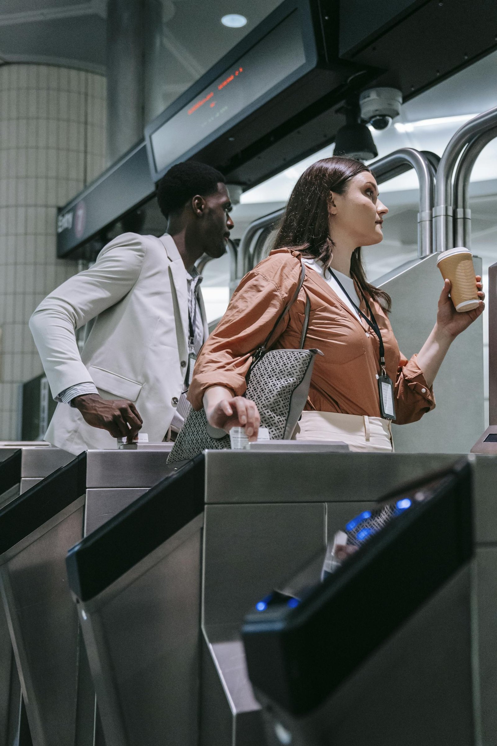 Black man and Caucasian woman rush through a subway turnstile holding a coffee and a laptop case.