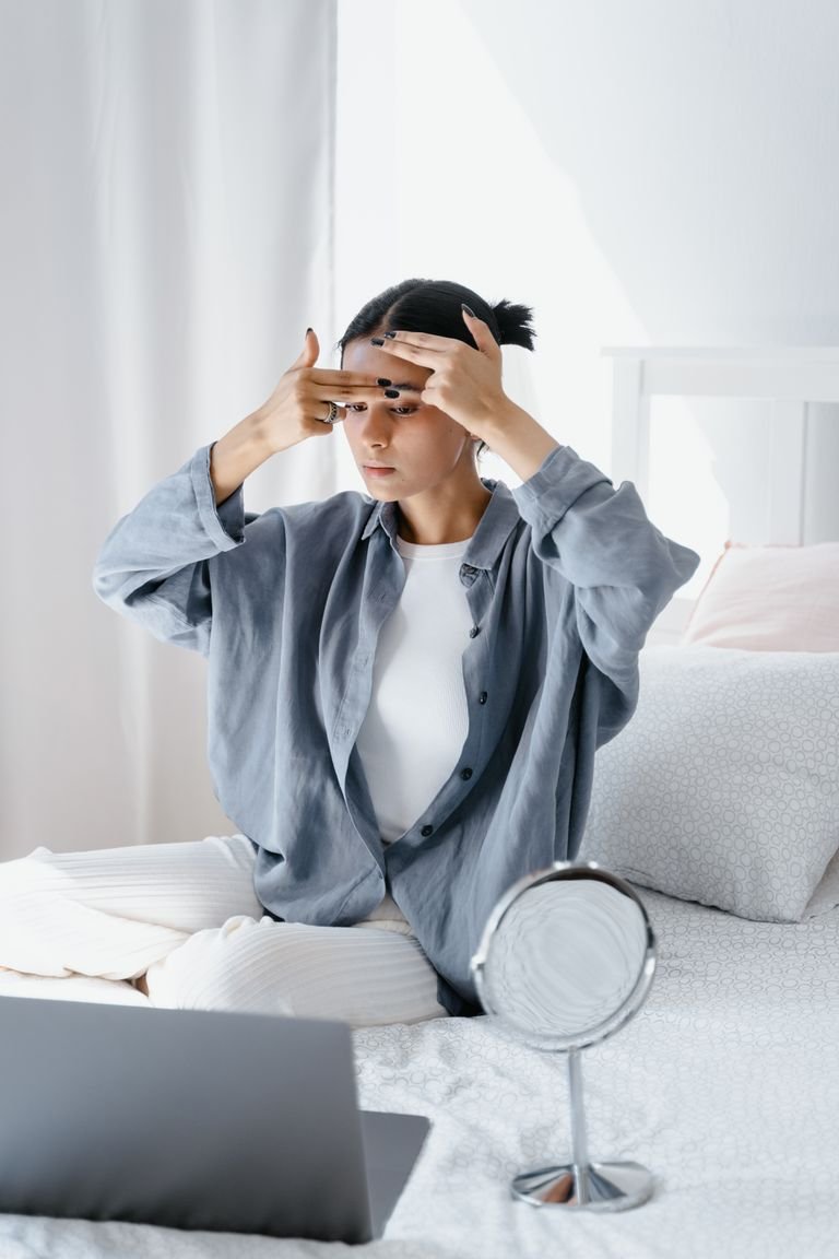 Young woman applying skincare at home with a mirror, focusing on forehead.